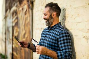 Portrait of modern businessman with beard holding credit card and mobile phone while standing in front of brick wall outdoor photo
