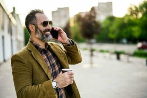 Portrait of modern businessman  with beard using mobile phone and drinking coffee while standing on the city street photo