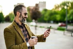 Portrait of modern businessman  with beard using mobile phone and drinking coffee while standing on the city street photo