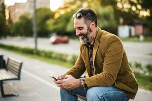 Modern businessman with beard using mobile phone while sitting on the bench in the city street photo