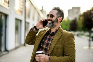 Portrait of modern businessman  with beard using mobile phone and drinking coffee while standing on the city street photo