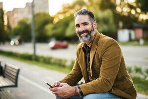 Modern businessman with beard using mobile phone while sitting on the bench in the city street photo