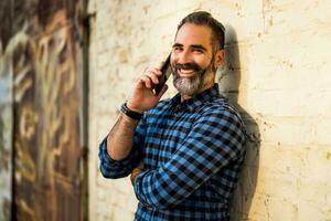 moderno empresario con barba utilizando móvil teléfono mientras en pie en frente de pared al aire libre foto