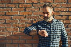 Modern businessman with beard looking at his watch while standing in front of brick wall outdoor photo