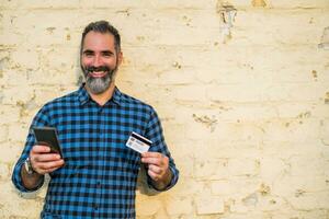 Portrait of modern businessman with beard holding credit card and mobile phone while standing in front of brick wall  outdoor photo