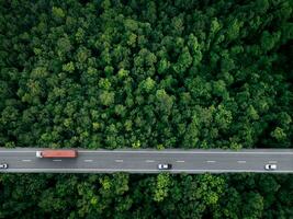 Aerial top view of car and truck driving on highway road in green forest. Sustainable transport. Drone view of hydrogen energy truck and electric vehicle driving on asphalt road through green forest. photo