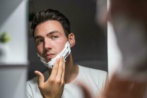 hombre aplicando afeitado crema mientras mirando él mismo en el espejo foto