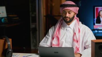 portrait de souriant arabe pigiste travail de maison, dactylographie sur le sien ordinateur portable, surfant le filet. milieu est homme dans traditionnel tenue navigation sur numérique appareil, Envoi en cours courriels à clients video