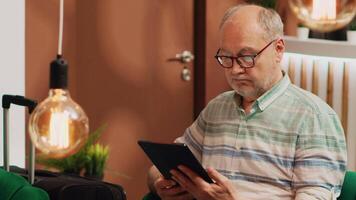 Older man tourist on lounge area sofa, surfing the web on digital tablet. Person holding device to browse internet waiting to see room at five star hotel on vacation, checks online reservation. video