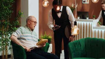 pensionerad man betalande för kopp av kaffe använder sig av kreditera kort i lobby, beställning förfriskning från hotell bar och framställning pos transaktion. bellboy concierge tjänande klient med dryck medan han är väntar. video