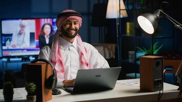 Portrait of happy Arab businessman working at home desk, typing on his laptop, solving tasks. Middle eastern man browsing on digital device, doing email communication in professional office setting video