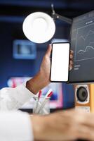 Detailed view of a male hand holding a mobile device with an isolated copyspace white screen in front of a desktop pc. Arab person displaying a blank chromakey mockup template on a smartphone. photo