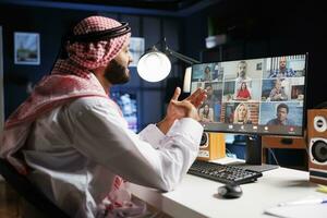 imagen exhibiendo un medio oriental hombre vistiendo tradicional ropa, Hablando con un grupo de personas en su computadora pantalla. un islámico chico conversando con su equipo en un conferencia llamar. foto