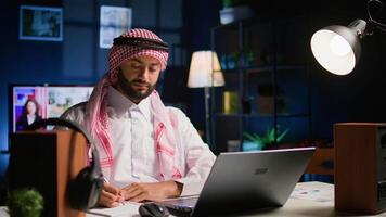 Portrait of happy arab teleworker remotely working in apartment writing on paper. Middle Eastern employee at home cross referencing information, transcribing data from laptop to notepad video