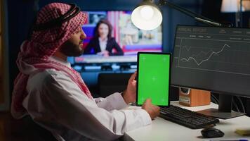 Remote worker at home holds chroma key tablet while looking at business annual report graphs on computer. Arab freelancer with mockup digital device in hand performing company tasks video