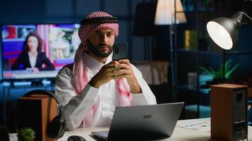 portret van glimlachen Arabisch freelancer genieten van vrije tijd Bij huis, typen Aan zijn smartphone, surfing de netto. gelukkig midden- oostelijk Mens in traditioneel kleding chatten met vrienden gebruik makend van mobiel telefoon video