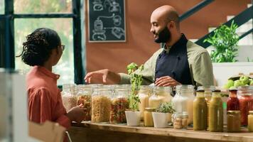 venditore regali massa prodotti nel barattoli, consigliare genere di bio biologico pasta per femmina cliente nel Locale drogheria negozio. giovane uomo mostrando massa prodotti nel riciclabile contenitori, salutare mangiare. video