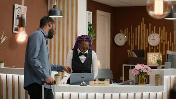 Person calls for service and room card at reception using front desk bell to talk to receptionist in lobby. African american hotel guest with trolley bags receiving access key for accommodation. photo