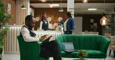 African american staff making online bookings on tablet in lobby, working at luxurious hotel and providing excellent concierge service for guests upon their arrival. Bellboy in uniform manage rooms. photo