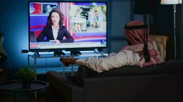 Arabic man lounged on cozy sofa, watching news broadcast while relaxing in apartment after hard day at work. Middle Eastern person chilling on couch, looking at TV channel showing current events video