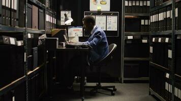 African american private detective walks into archive room with a laptop bag and starts working crime investigation. Male investigator sits at desk with wireless computer, checking suspect case files. photo