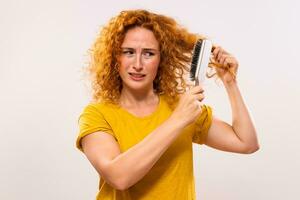 Angry ginger woman holding hairbrush and combing her curly hair photo