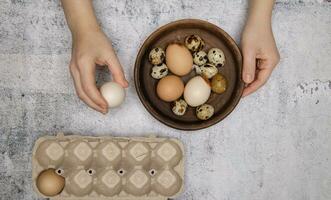 manos de invisible persona llevar Fresco pollo y codorniz huevos desde marrón arcilla plato a huevo bandeja. gris mesa. parte superior vista. alto calidad foto