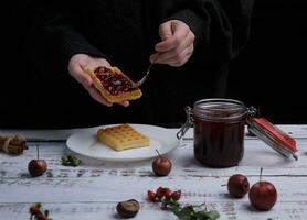 hands hold waffles and a spoon and spread them with strawberry jam from a jar. selective focus photo