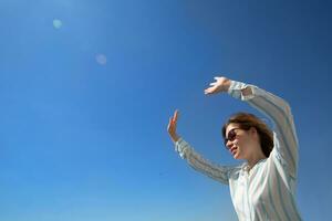 happy blonde in glasses and a light shirt waving her hand to someone on a background of blue sky. High quality photo