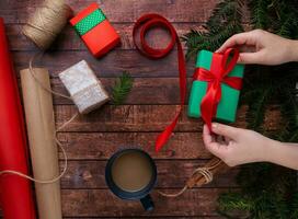 hands are packing Christmas gifts and tying green box with red ribbon. There is cup of coffee on the table. photo