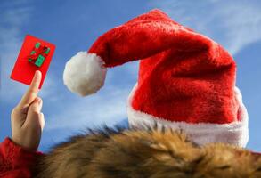 mujer en Papa Noel sombrero participación un regalo tarjeta o crédito tarjeta representando un regalo caja en contra el azul cielo. vistiendo un Papa Noel sombrero y un rojo chaqueta. regalo o descuento concepto.selectivo enfocar. foto