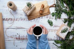 hands in blue sweater hold white cup of coffee on an old wooden work table with handmade gifts and branches of Christmas tree.Flat Lay.Top view photo