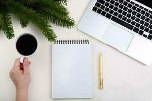 hand holds a cup of coffee near a laptop, pen, notepad and branches of a Christmas tree. cropped image. High quality photo