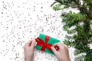 Hands hold one Christmas gift with red ribbon on the white table with Christmas tree nearby . Christmas wrapping photo