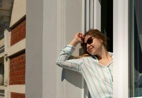 Happy blonde woman in sunglasses sits on windowsill and smiles at the sun. High quality photo