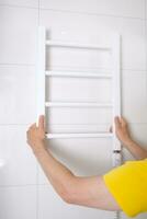 Renovation in the bathroom. A plumber in a yellow shirt installs a heated towel rail. Interior walls with light tiles. High quality photo