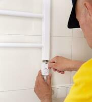 Repair in the bathroom. Male hands of a worker with a screwdriver. Plumber sets a heated towel rail. The interior of the walls with light tiles. photo