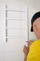 Repair in the bathroom. Male hands of a worker with a screwdriver. Plumber sets a heated towel rail. The interior of the walls with light tiles. photo