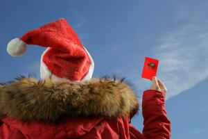 mujer en Papa Noel sombrero participación un regalo tarjeta o crédito tarjeta representando un regalo caja en contra el azul cielo. vistiendo un Papa Noel sombrero y un rojo chaqueta. regalo o descuento concepto.selectivo atención foto