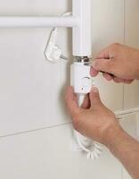 Repair in the bathroom. Male hands of a worker with a screwdriver. Plumber sets a heated towel rail. The interior of the walls with light tiles. photo