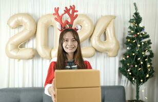asiático mujer participación un regalo caja tener divertido celebrando Navidad 2024. Decorar y Decorar el habitación con un Navidad árbol y 2024 globos foto