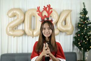 asiático mujer vestido como un chabola aplaudir tu manos felizmente en anticipación de celebrando Navidad 2024. Decorar y Decorar el habitación con Navidad arboles y globos 2024. foto