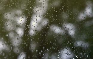 A photo of rain drops on the window glass with a blurred view of the blossoming green trees. Abstract image showing cloudy and rainy weather conditions