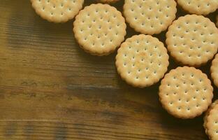 A round sandwich cookie with coconut filling lies in large quantities on a brown wooden surface. Photo of edible treats on a wooden background with copy space