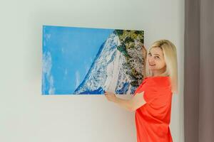 A happy young blonde woman is holding a large wall canvas at home photo