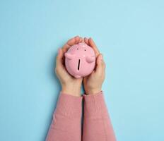 Female hands hold a pink ceramic piggy bank against a blue background, symbolizing the concept of saving money photo