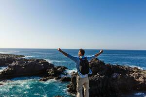 A young man on the rocks and looking at the ocean while a big wave is breaking in front of him photo
