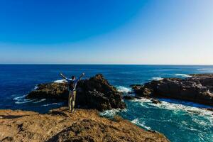 A young man on the rocks and looking at the ocean while a big wave is breaking in front of him photo