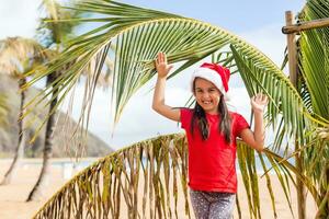 pequeño niña en Papa Noel sombrero en pie siguiente a palma árbol en playa foto