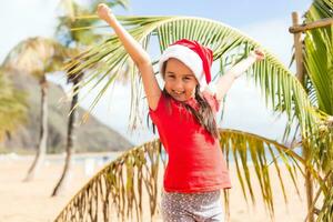 Little girl in santa hat standing next to palm tree on beach photo